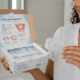 A woman smiling while holding an over-the-counter teeth whitening kit in a bathroom setting.